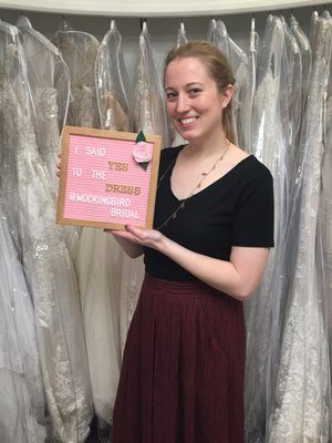 A bride holding the I-said-yes-at-Mockingbird-Bridal sign by the gown racks